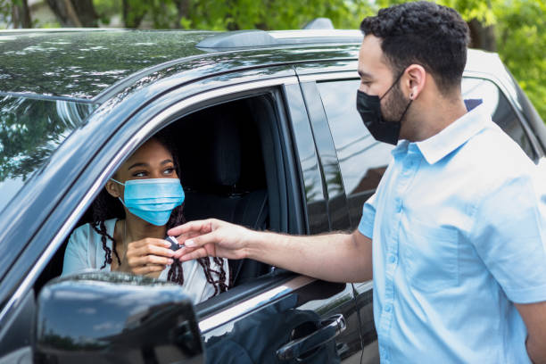 Sitting in her new car, the mid adult woman receives the keys for the car from the mid adult salesman.  They both wear masks because of COVID-19.